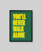 A framed typographic wall art print with the phrase 'YOU'LL NEVER WALK ALONE' in bold yellow letters on a dark green background, shown against a light gray wall.