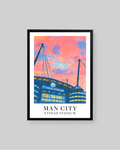 A framed art print of Etihad Stadium in Manchester, featuring a colorful, textured sketch of the stadium under a vibrant pink and orange sky, with 'MAN CITY ETIHAD STADIUM' printed at the bottom. The print is framed in black with a white mat, set against a solid gray background.